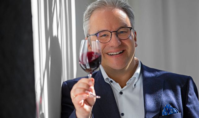 Ein Mann im Anzug lehnt an einer Wand und hält in seiner einen Hand ein Glas Rotwein. Er lächelt in die Kamera.