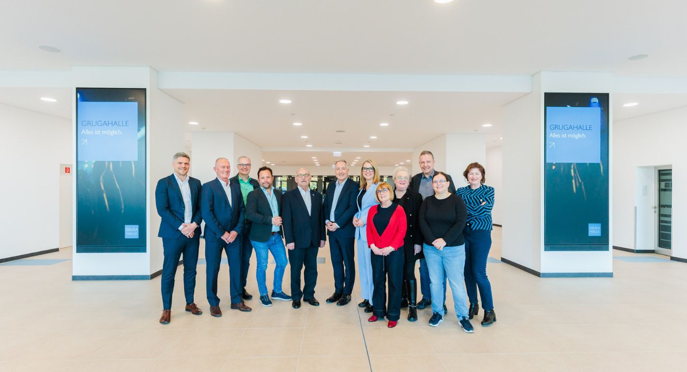 Gruppenfoto von 12 Personen in einem modernen, hell beleuchteten Foyer der Grugahalle in Essen. Zwei digitale Tafeln mit dem Schriftzug 'Grugahalle – Alles ist möglich.' sind im Hintergrund zu sehen.