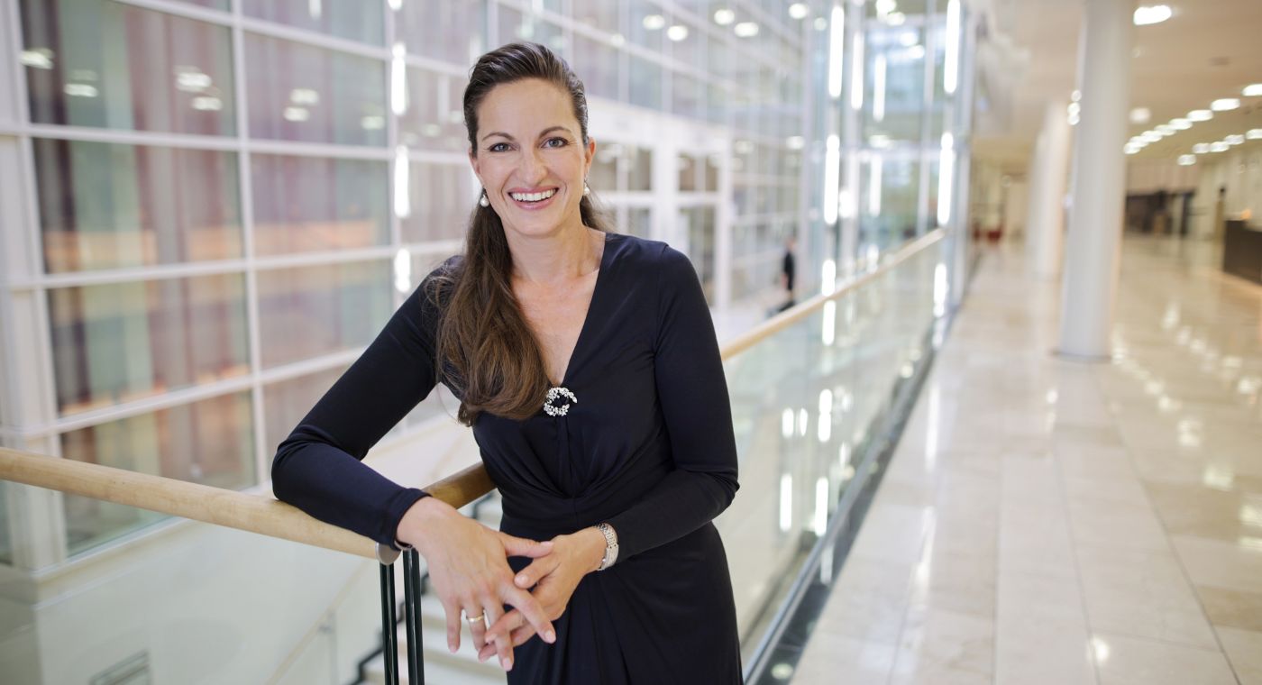 Eine Frau mit langen braunen Haaren und in elegantem, dunkelblauem Kleid steht lächelnd in einem modernen, hellen Gebäude.