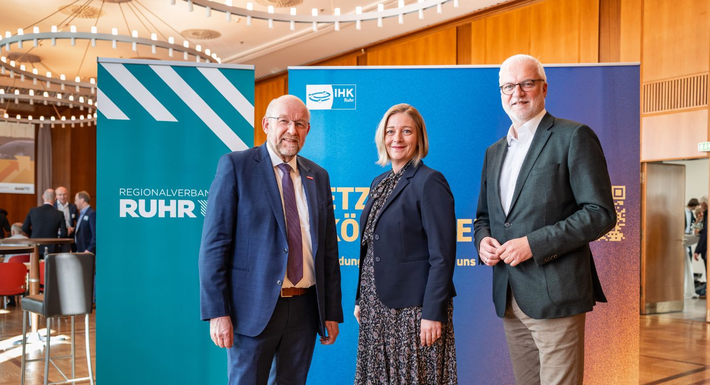 Zwei Männer und eine Frau stehen vor Roll-Ups des Regionalverbandes Ruhr und der IHK Ruhr bei einer Veranstaltung in einem Konferenzraum.
