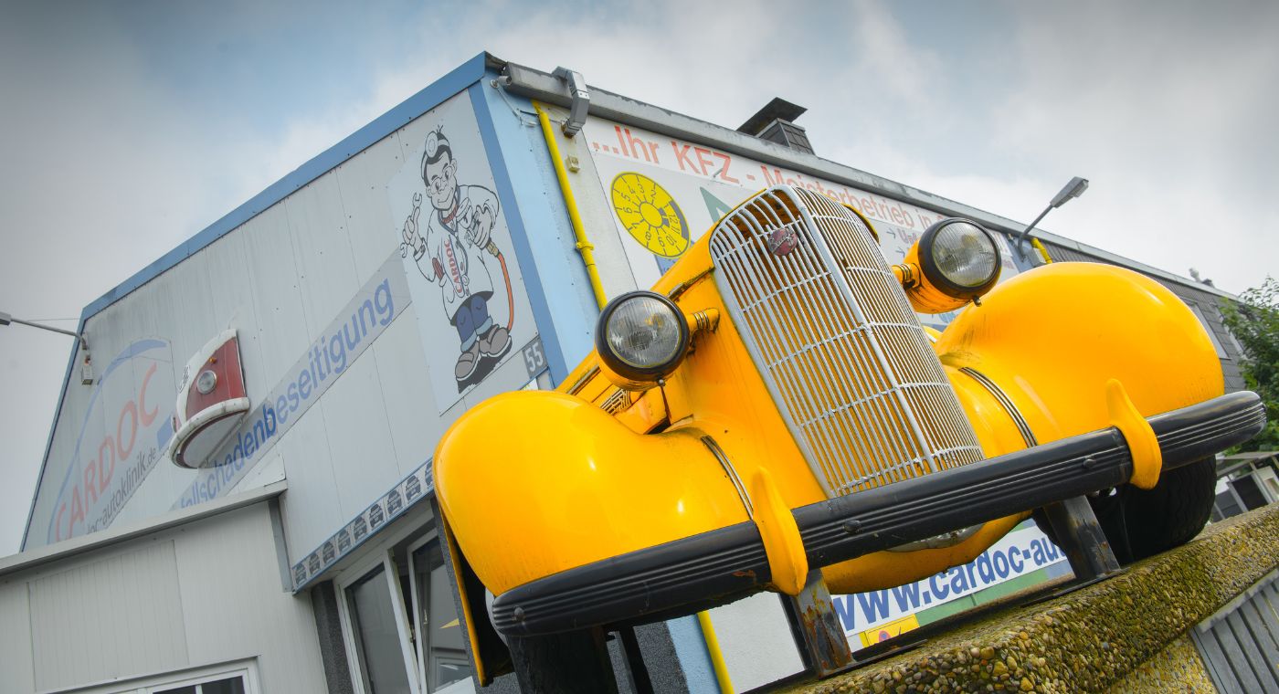 Ein auffällig gelber Oldtimer ragt als dekoratives Element aus der Fassade einer Autowerkstatt. Im Hintergrund ist das Gebäude mit dem Logo und der Beschriftung der Firma „CARDOC“ sowie einer Comicfigur eines Autodoktors zu sehen.