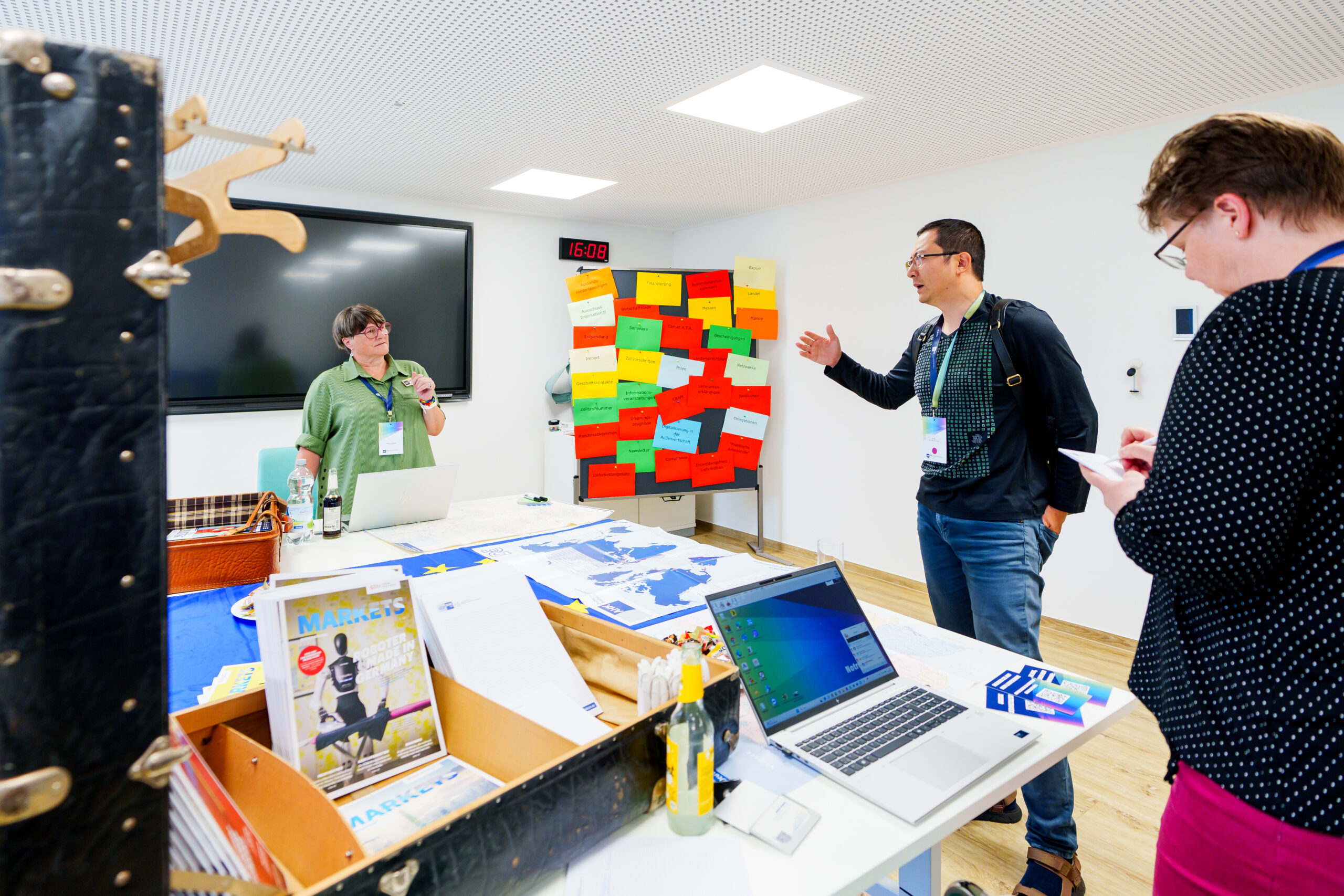 Drei Personen stehen in einem Raum und unterhalten sich. Auf dem Tisch steht ein Laptop. Im Hintergrund ist eine Flipchart mit Notizzetteln.