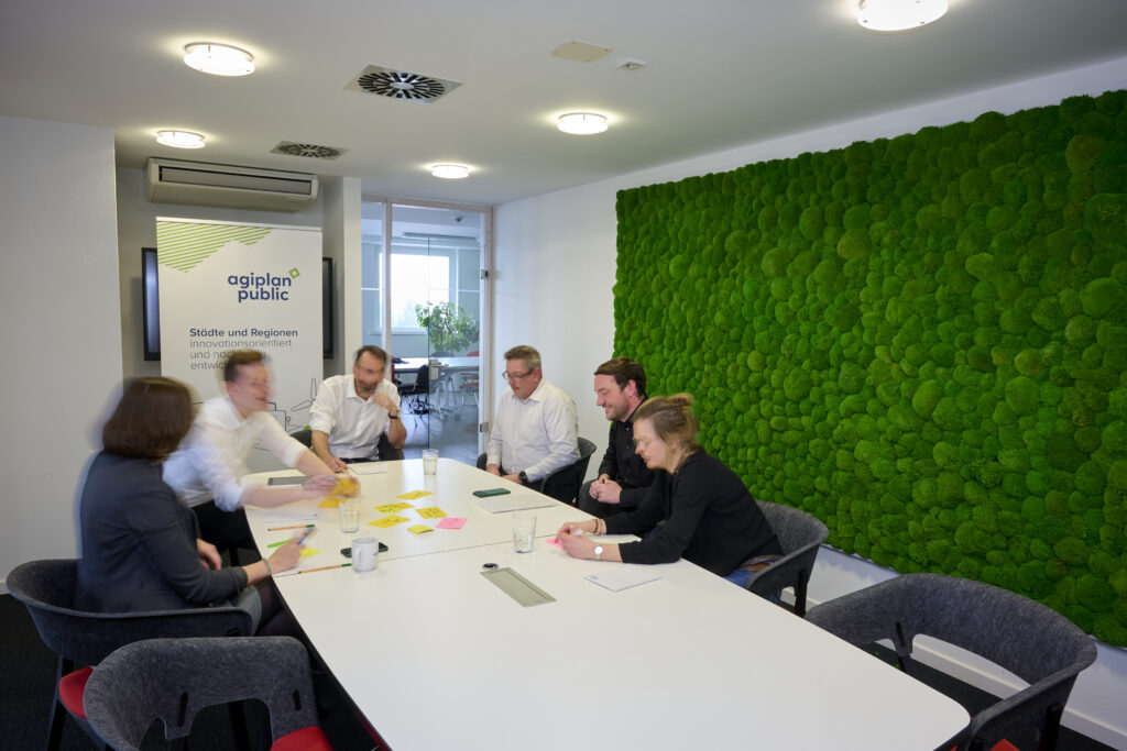 Sechs Personen sitzen am Tisch, unterhalten sich und arbeiten zusammen. Auf dem Tisch liegen Notizzettel und Stifte.