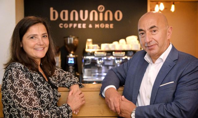 Ein Mann und eine Frau sitzen in einem Ladenlokal und lächeln. Im Hintergrund steht eine Kaffeemaschine.