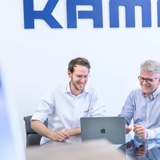 Zwei Männer sitzen an einem Tisch und schauen auf ein Notebook. Sie unterhalten sich und lächeln.