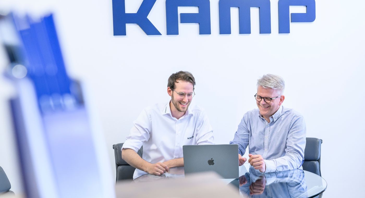 Zwei Männer sitzen an einem Tisch und schauen auf ein Notebook. Sie unterhalten sich und lächeln.