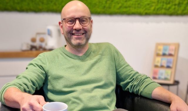 Ein Mann sitzt auf einem Sessel und hält eine Kaffeetasse. Er lächelt in die Kamera.