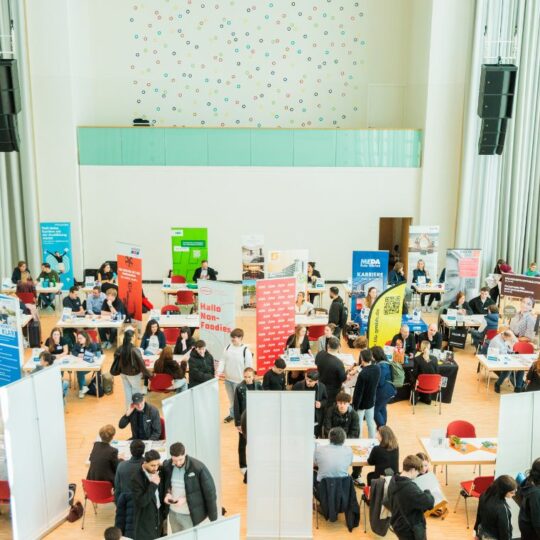 Ein großer Saal mit mehreren kleinen Ständen von Unternehmen. Drum herum laufen junge Menschen.