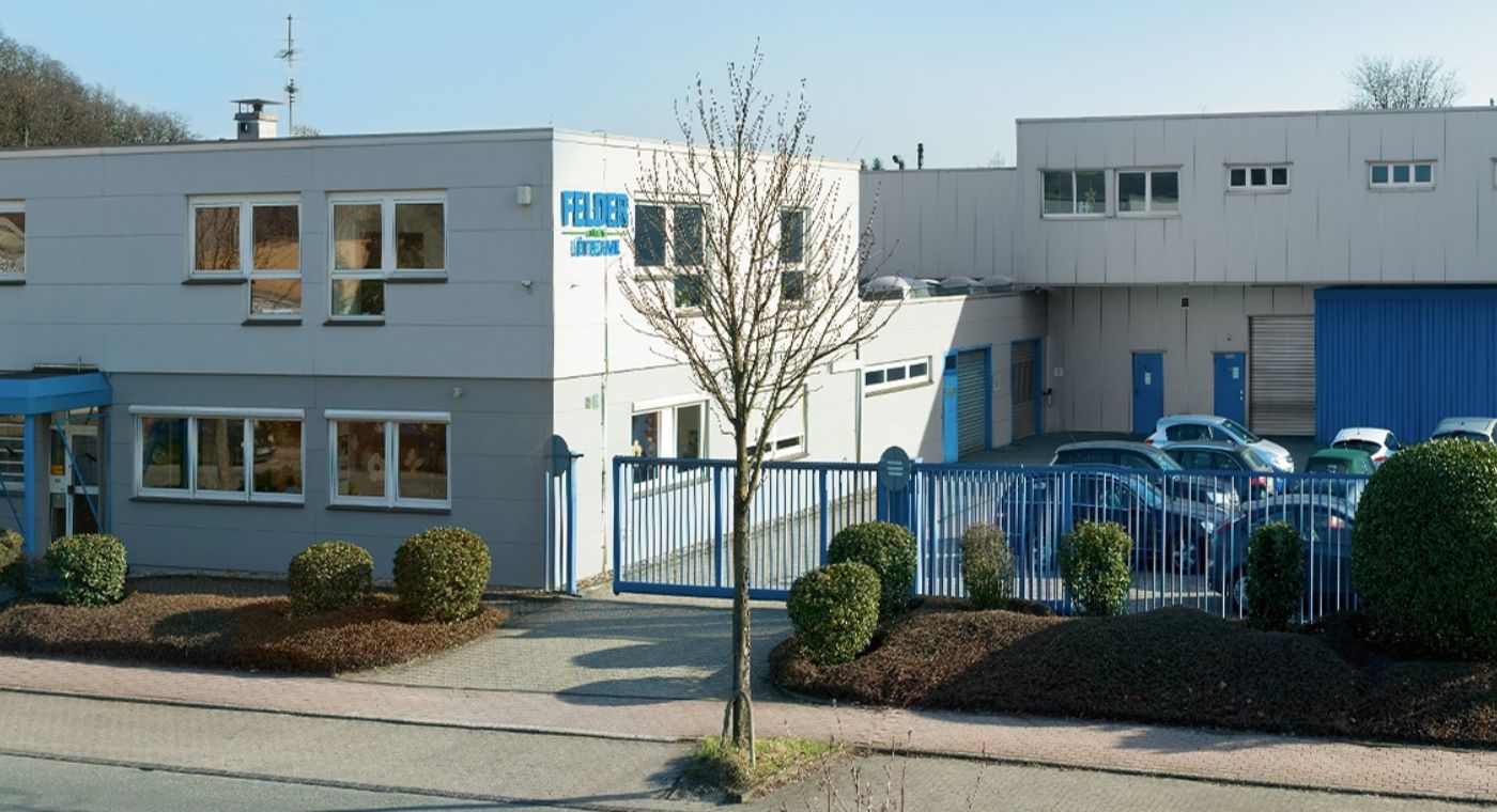 Links das Gebäude der FELDER GmbH. Rechts ein Parkpkatz mit Autos. Vor dem Gebäude ist eine Straße, Büsche und drei Bäume ohne Blätter.