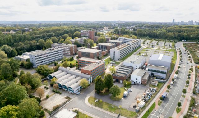 Luftaufnahme eines weitläufigen Campus mit mehreren modernen und älteren Gebäuden, umgeben von vielen Bäumen und Grünflächen. Im Vordergrund befinden sich Industrie- und Bürogebäude mit Flachdächern sowie eine Straße.