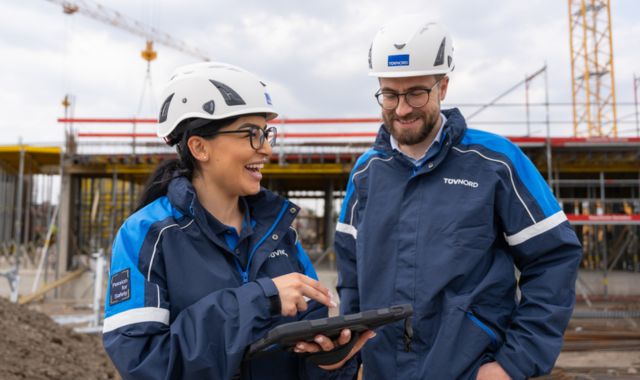Eine Frau und ein Mann stehen auf einer Baustelle. Die Frau hält ein Tablet in der Hand, beide lächeln. Im Hintergrund sind zwei Krane und ein Gebäude, das gerade gebaut wird.