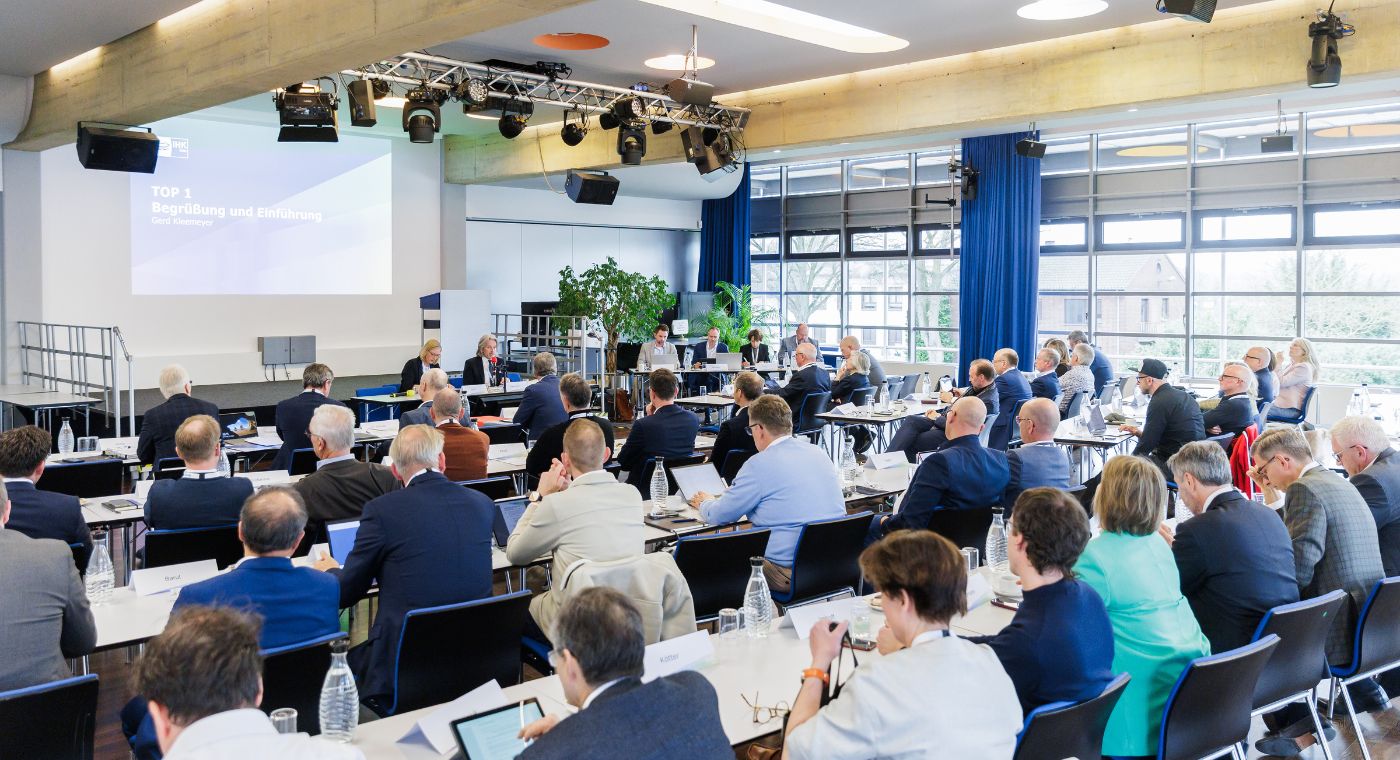 In einem großen Saal sitzen viele Personen und hören zwei Menschen vorne zu. Im Hintergrund läuft eine Präsentation.