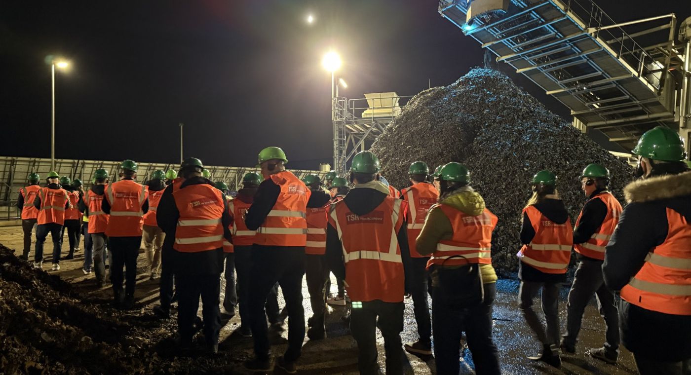 Mehrere Personen laufen hintereinander draußen über eine Baustelle. Draußen ist es dunkel. Alle haben einen Helm auf und Warnwesten an.