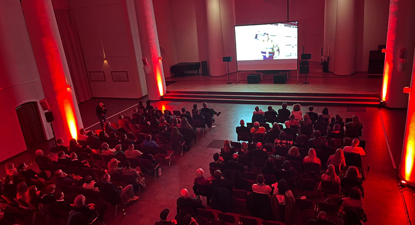 Viele Menschen sitzen in einem großen Saal. Vorne läuft ein Unternehmensfilm.