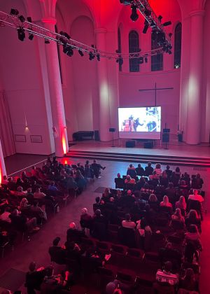 Publikum sitzt in einem großen Saal. Auf einer kleinen Bühne läuft ein Film.