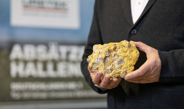 Person im Anzug hält ein gelblich-graues Gesteinsstück mit sichtbaren Bruchstrukturen in den Händen.