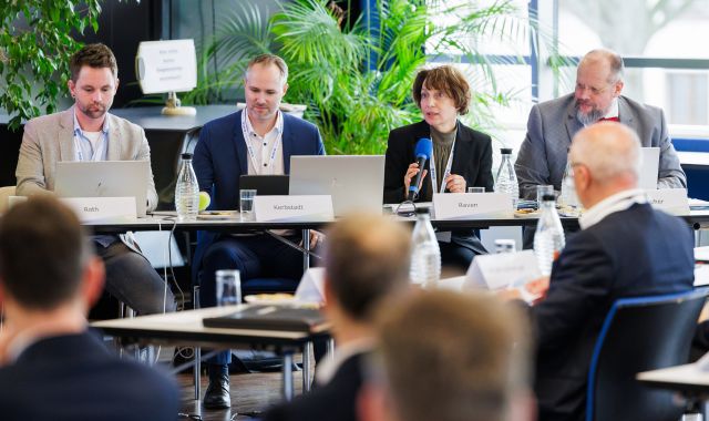 Vier Personen sitzen an einem Tisch. Eine Frau hält ein Mikrophon und redet.