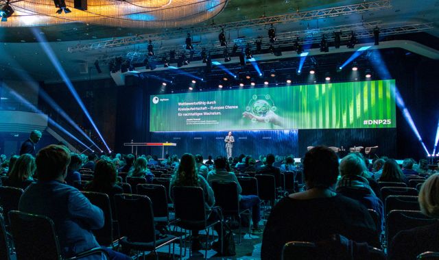 Viele Gäste sitzen in einem großen Saal und schauen nach vorne. Auf einer Bühne steht ein Mann und trägt etwas vor.