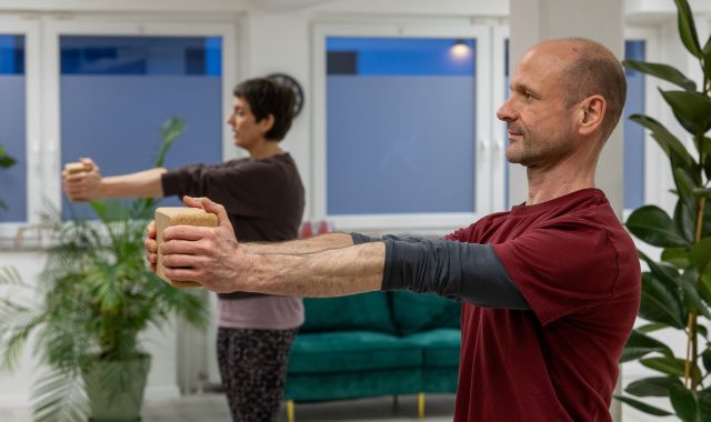 Zwei Personen stehen in einem Yoga-Studio, strecken ihre Arme aus.