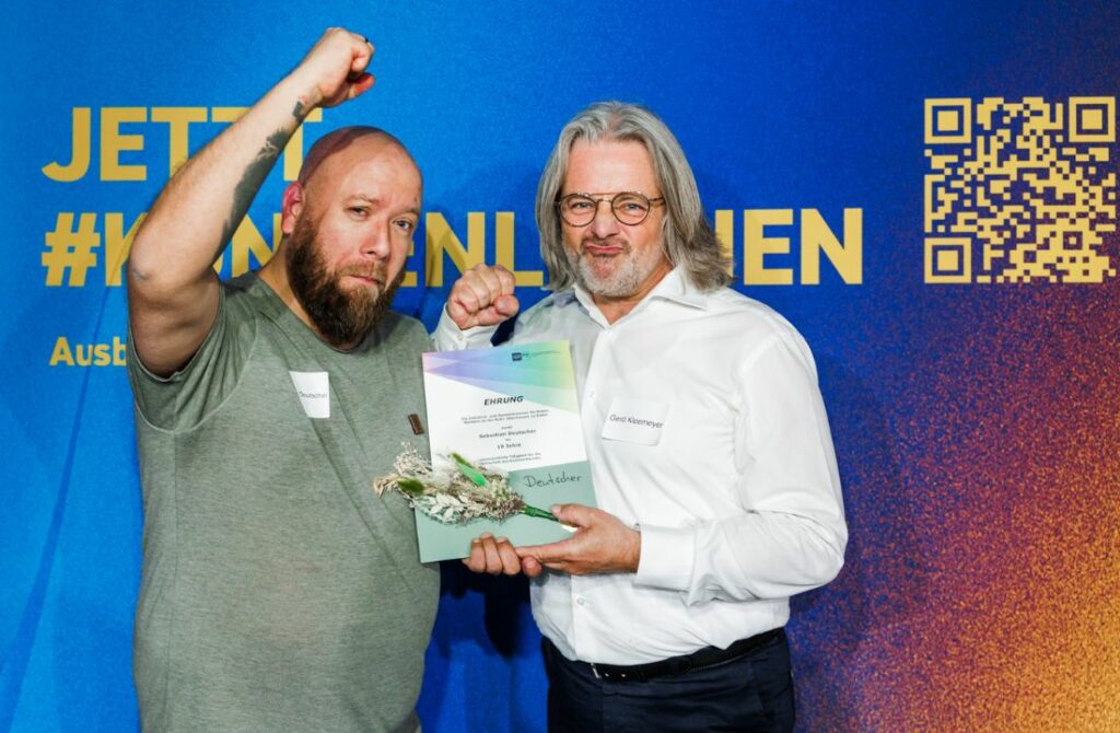 Zwei Männer halten eine Urkunde und schauen in die Kamera.