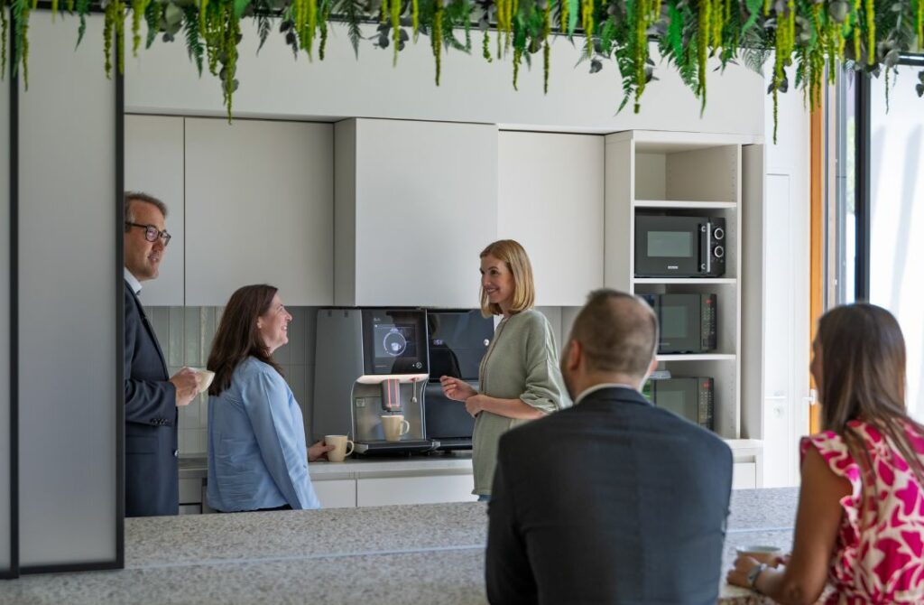 5 Personen stehen in der Küche und unterhalten sich. Eine Frau bedient eine Kaffeemaschine.