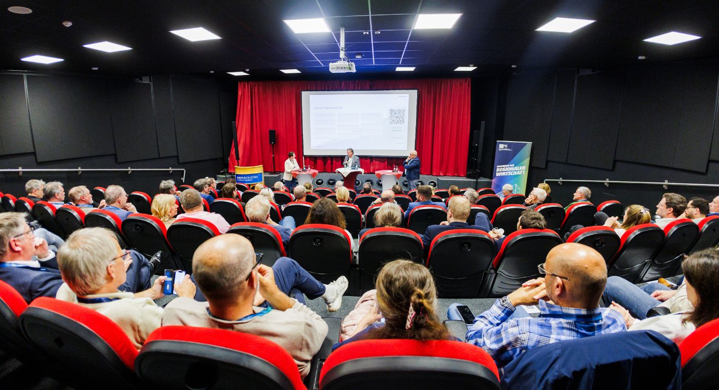 Mehrere Menschen sitzen in einem Saal. Auf einer Bühne stehen drei Menschen und sprechen. Im Hintergrund läuft eine Präsentation.