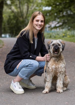 Eine Frau lächelt in die Kamera. Neben ihr sitzt ein Hund und schaut auch in die Kamera. Im Hintergrund ist ein Park.