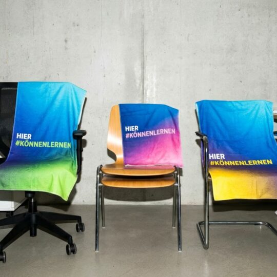 Auf drei Stühlen liegen Handtücher mit der Aufschrift "Hier #könnenlernen"