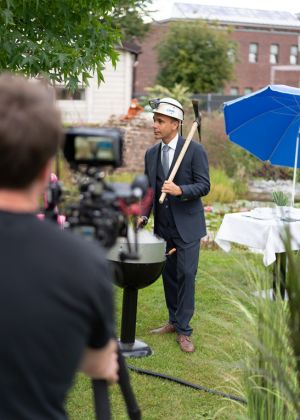 Ein Mann im Anzug steht im Freien auf einer Grünfläche und trägt einen weißen Bauhelm. In der Hand hält er eine Spitzhacke und spricht vor einer Kamera, die im Vordergrund zu sehen ist. Neben ihm stehen ein Grill und ein gedeckter Tisch mit blauem Sonnenschirm.