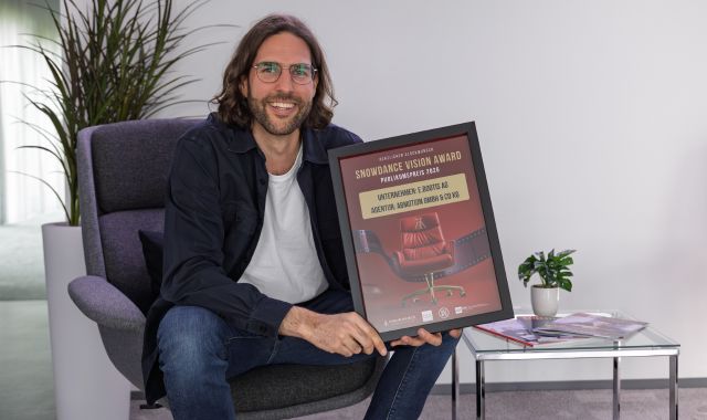 Ein Mann sitzt in einem Sessel in einem Büro und hält eine gerahmte Auszeichnung in die Kamera. Auf der Urkunde ist der „Snowdance Vision Award Publikumspreis 2026“ für die e.bootis AG zu sehen. Neben ihm stehen ein kleiner Glastisch mit Broschüren und eine Zimmerpflanze.