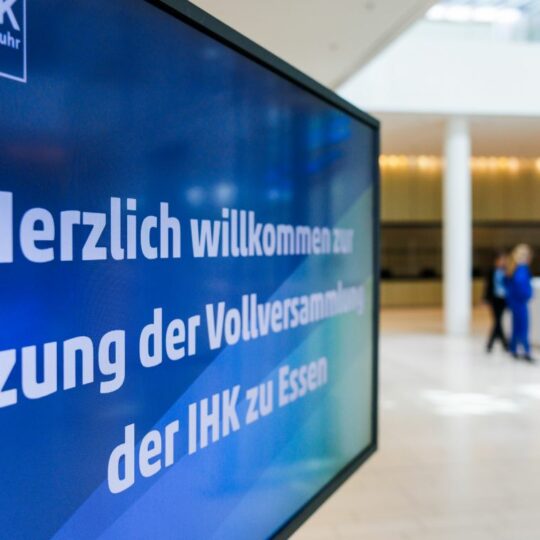 Auf einem Monitor läuft eine Präsenation: "Herzlich willkommen zur Sitzung der Vollversammlung 2026"
