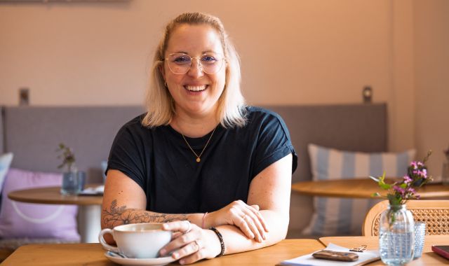 Eine Frau sitzt an einem Tisch und lächelt in die Kamera. Auf dem Tisch steht eine Blume und eine Kaffeetasse.