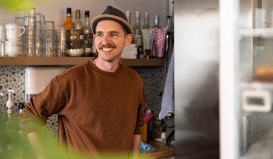 Ein Mann hat einen Hut auf, steht in einem Café und lächelt.