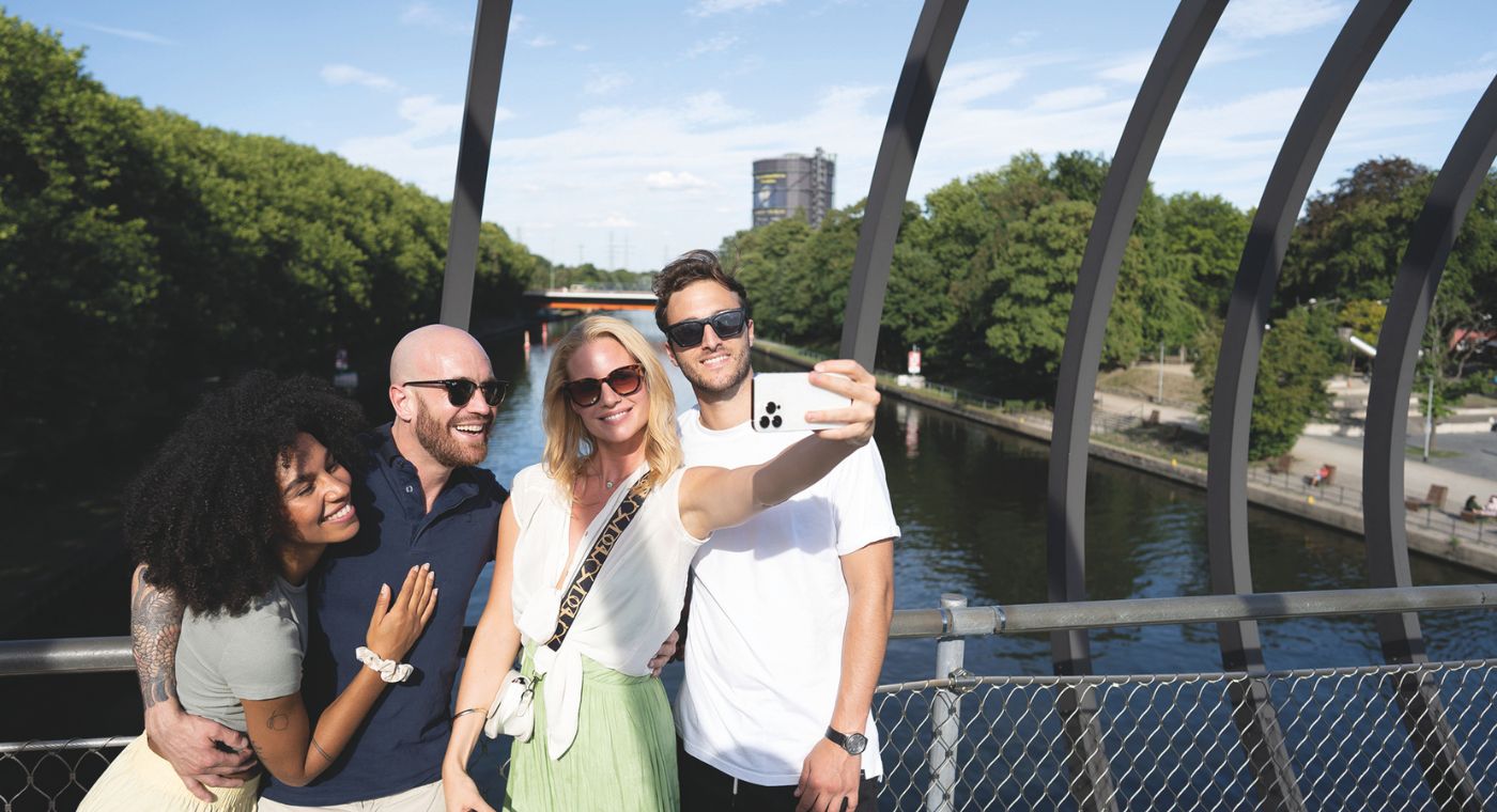 Vier Menschen stehen auf einer Brücke. Eine von ihnen hält ein Handy in der Hand und macht mit den anderen Drei ein Foto. Sie lächeln in die Kamera.