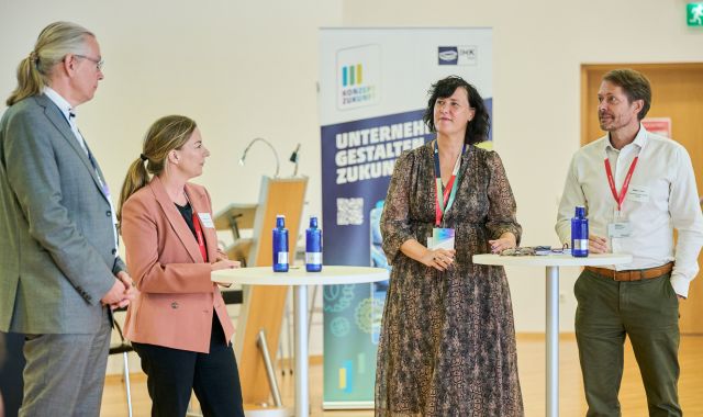 Vier Menschen stehen um zwei Stehtische herum und schauen sich an.