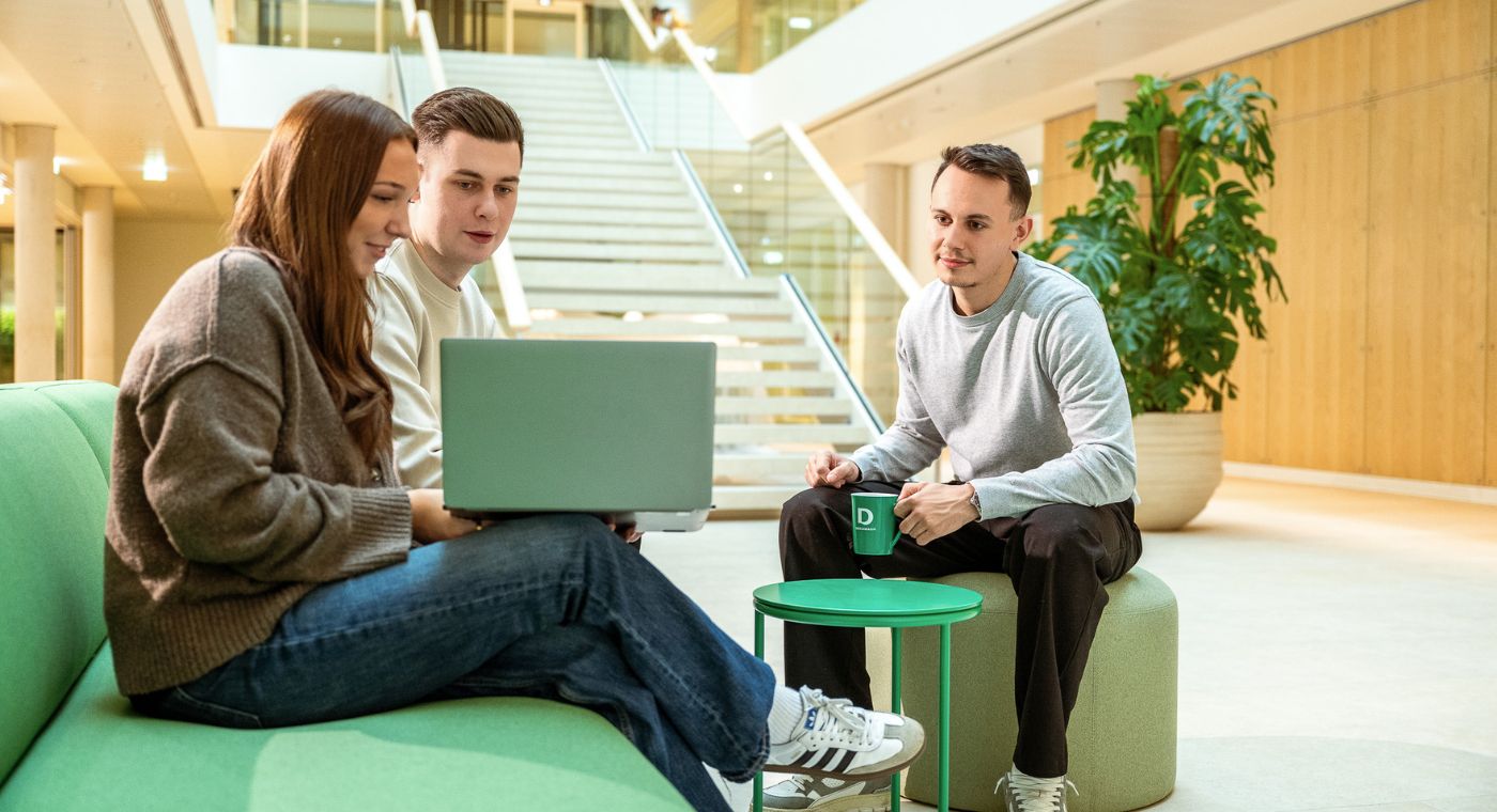 Drei junge Menschen sitzen in einem Gebäude. Ein Mädchen zeigt den zwei Jungs etwas auf einem Laptop.