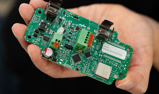 Eine Person hält eine grüne, unbestückte bzw. offenliegende Elektronikplatine in der Hand. Auf der Platine sind verschiedene Mikrochips, Anschlüsse, Klemmen und weitere elektronische Bauteile zu sehen.