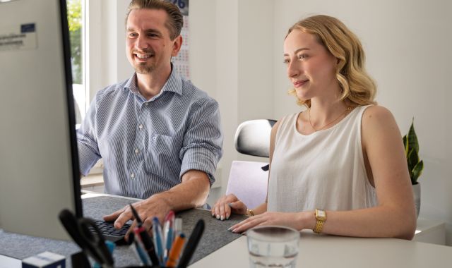 Ein Mann und eine Frau schauen auf einen Bildschirm. Sie sitzen an einem Tisch.